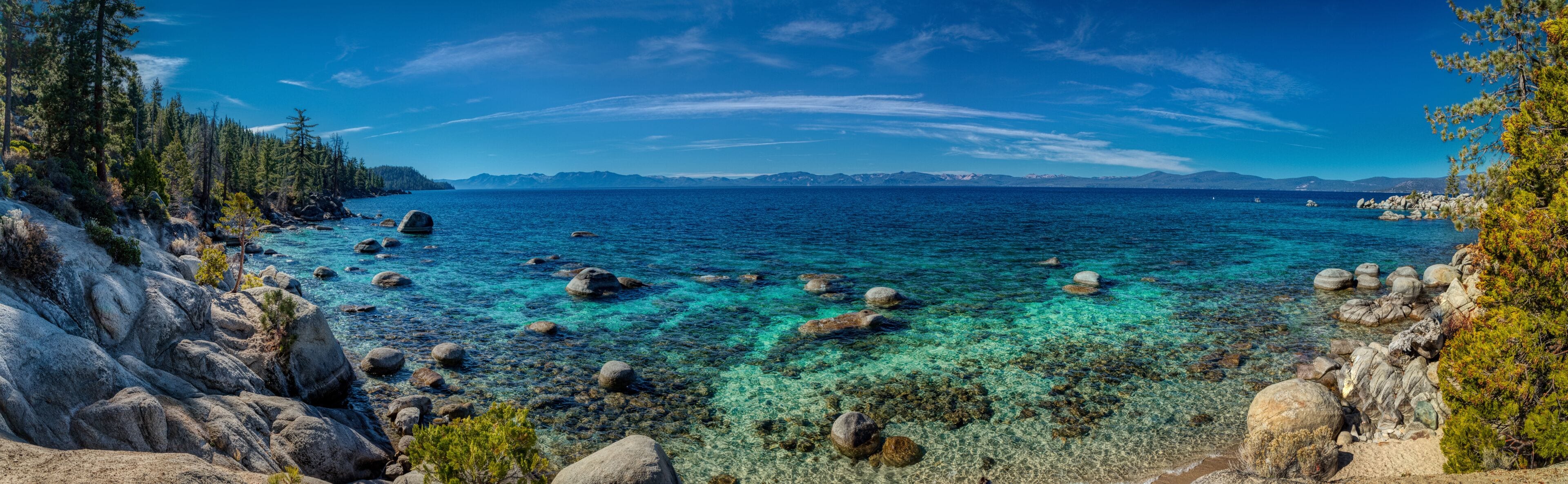 A clear coastal landscape with rocky shores, turquoise waters, and distant mountains under a blue sky.