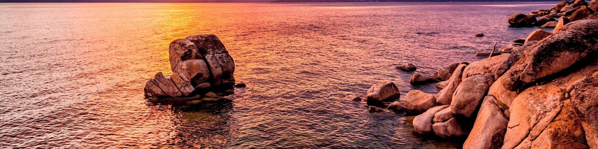 Un atardecer sobre un lago de aguas cristalinas, rodeado de grandes rocas y montañas al fondo.