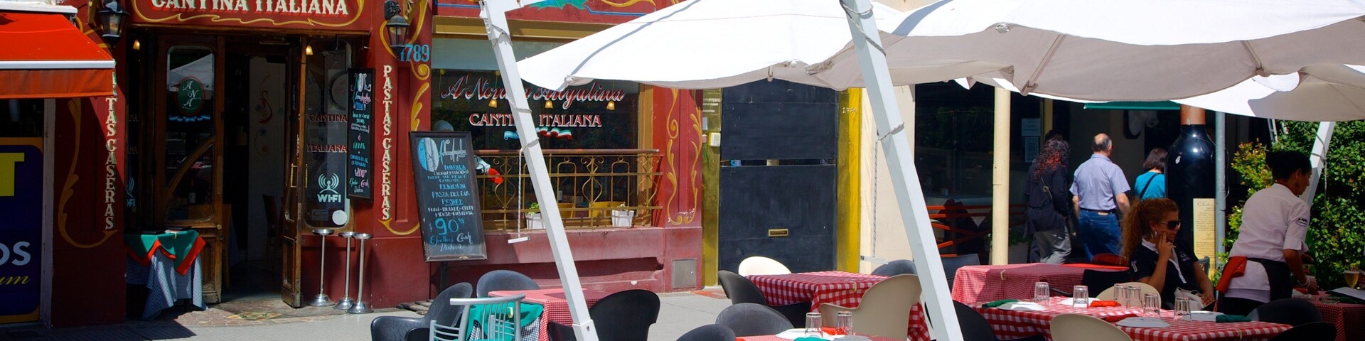 Mesas y sillas al aire libre con manteles a cuadros rojos y blancos, una fachada con un reloj y una bandera.