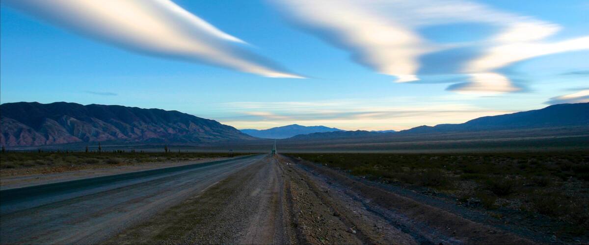 Un chemin de terre qui serpente à travers un paysage désertique, avec des montagnes à l’horizon et un ciel dégagé.