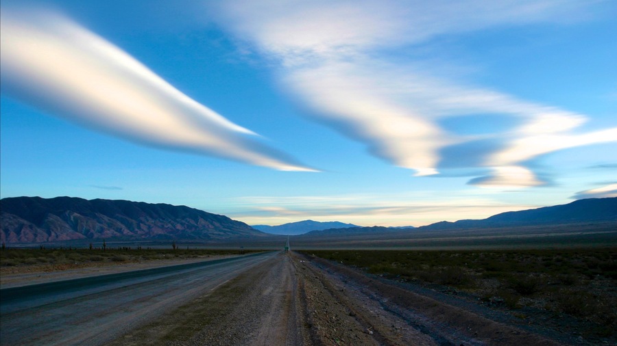 A dirt road stretching through a desert landscape with mountains in the distance and a clear sky.