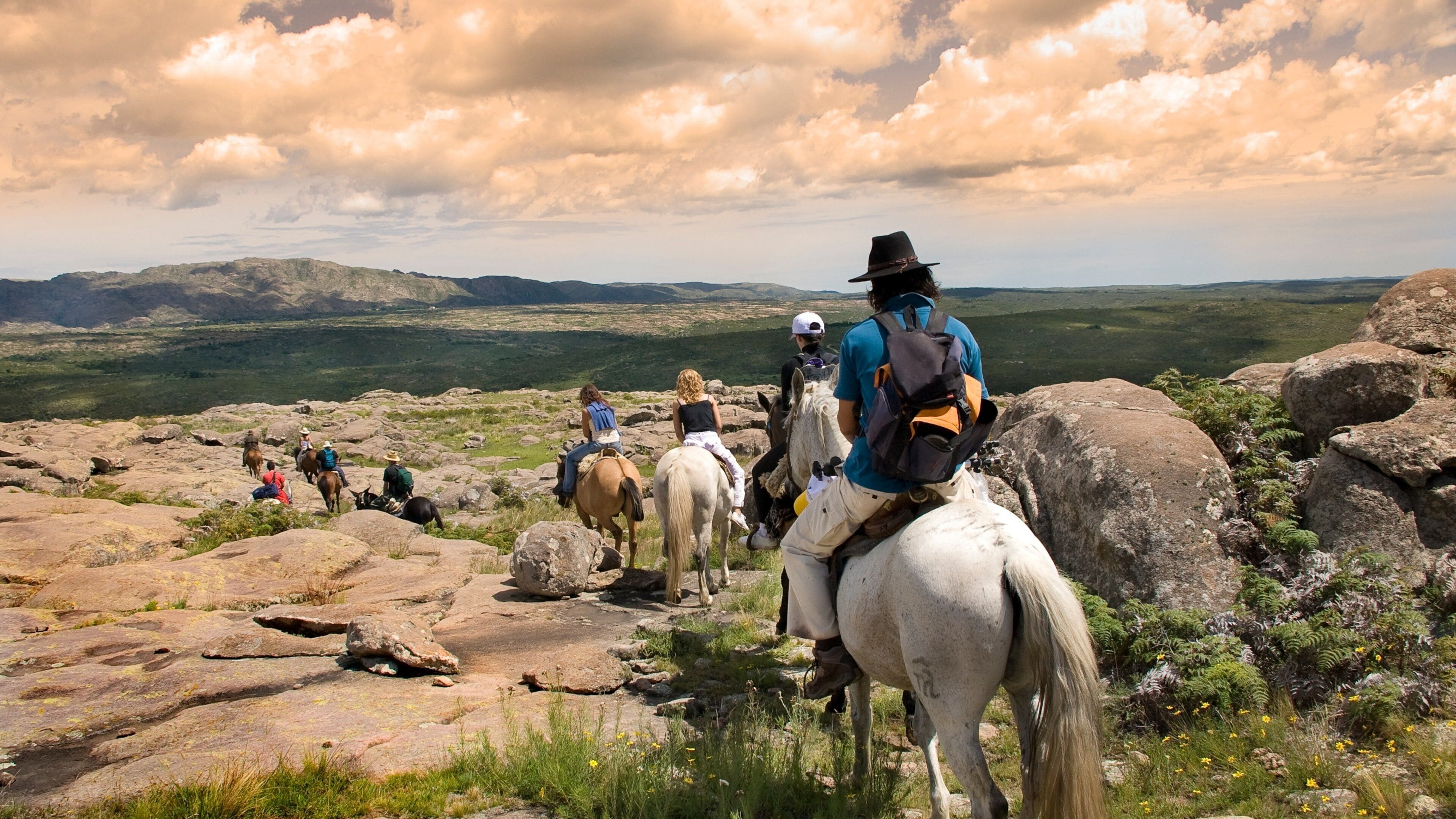 A group of people riding horses across a rocky landscape with mountains in the background.
