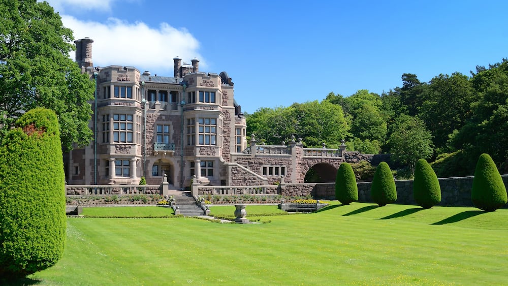 A large, historic stone building with a formal garden and trimmed hedges.