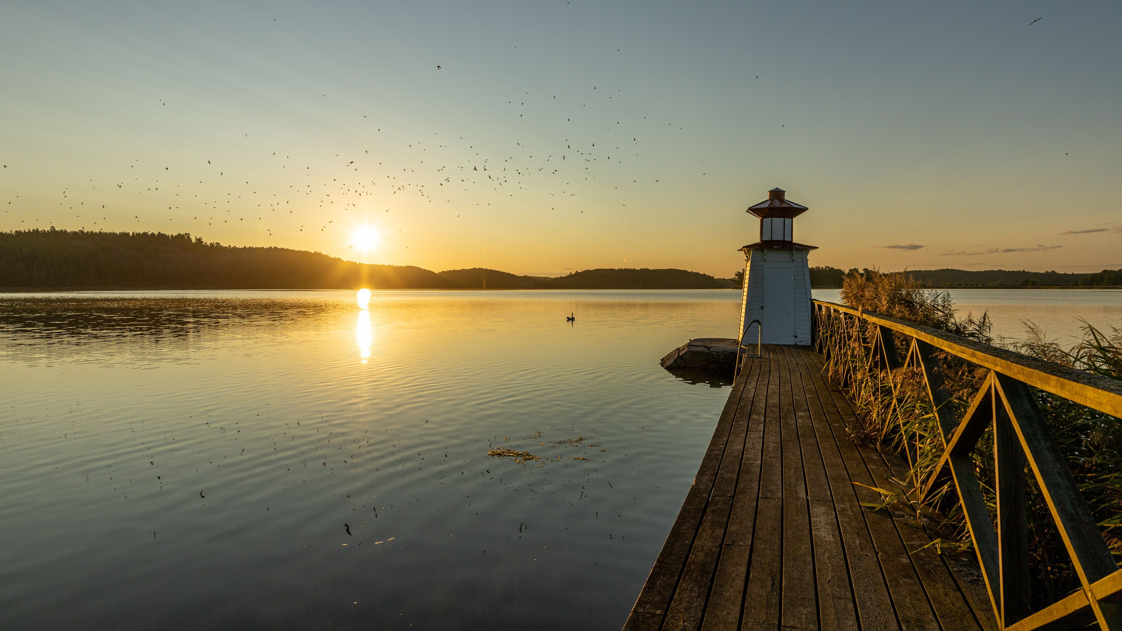 A wooden pier extending into calm water at sunset with a lighthouse in the background.