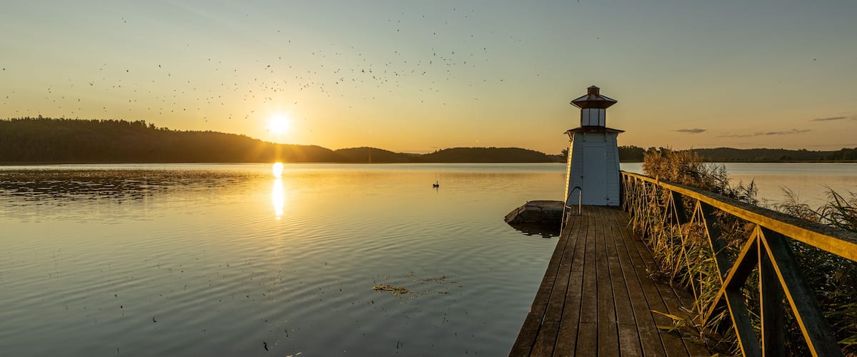 A wooden pier extending into calm water at sunset with a lighthouse in the background.