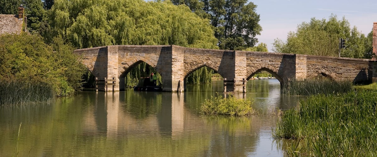 A stone bridge over a calm river, surrounded by lush greenery and trees.