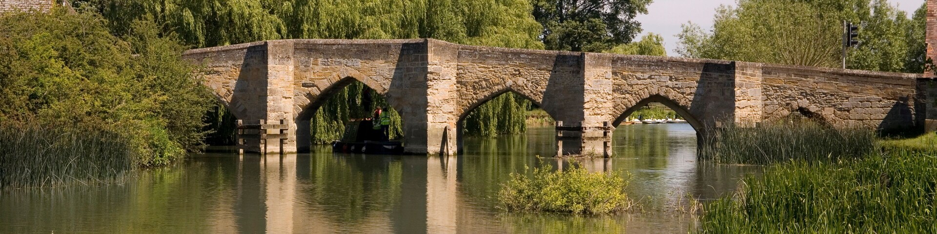 A stone bridge over a calm river, surrounded by lush greenery and trees.