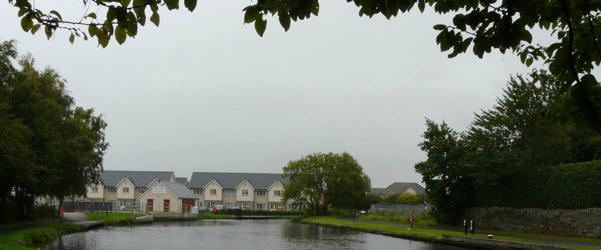 A calm river with a bridge, surrounded by greenery and a row of houses.