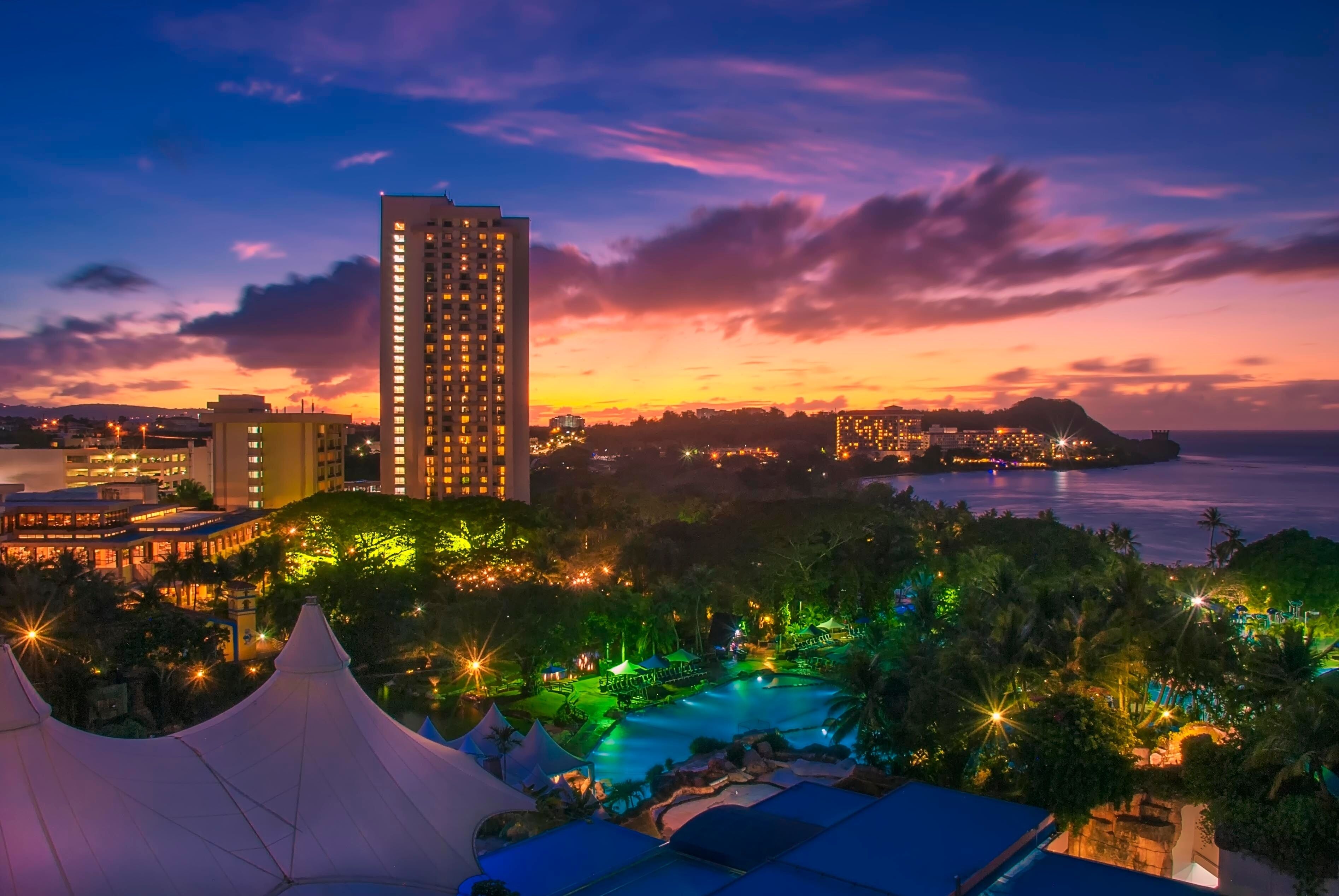 A resort with a swimming pool, a tall hotel, and a sunset over the ocean.