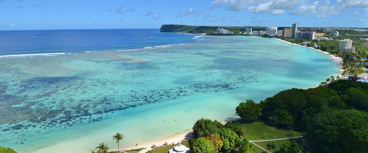 Un paisaje urbano costero con una playa, aguas turquesas cristalinas y un telón de fondo de edificios y vegetación.