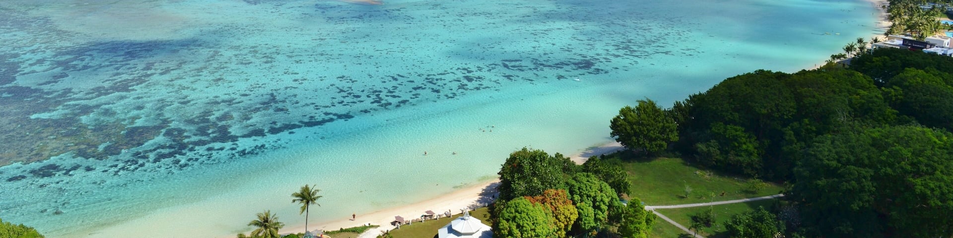 A coastal cityscape with a beach, clear turquoise waters, and a backdrop of buildings and greenery.