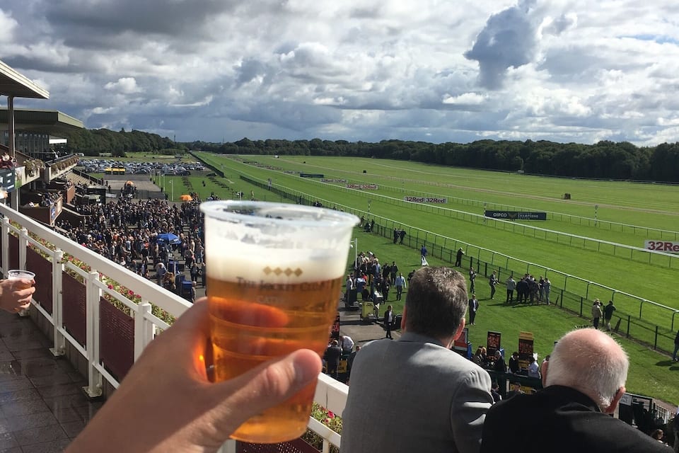 Una persona sosteniendo una jarra de cerveza con vistas a una pista de carreras y espectadores.