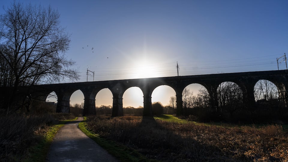 A stone viaduct with multiple arches over a rural road, surrounded by grass and trees, with the sun setting in the background.