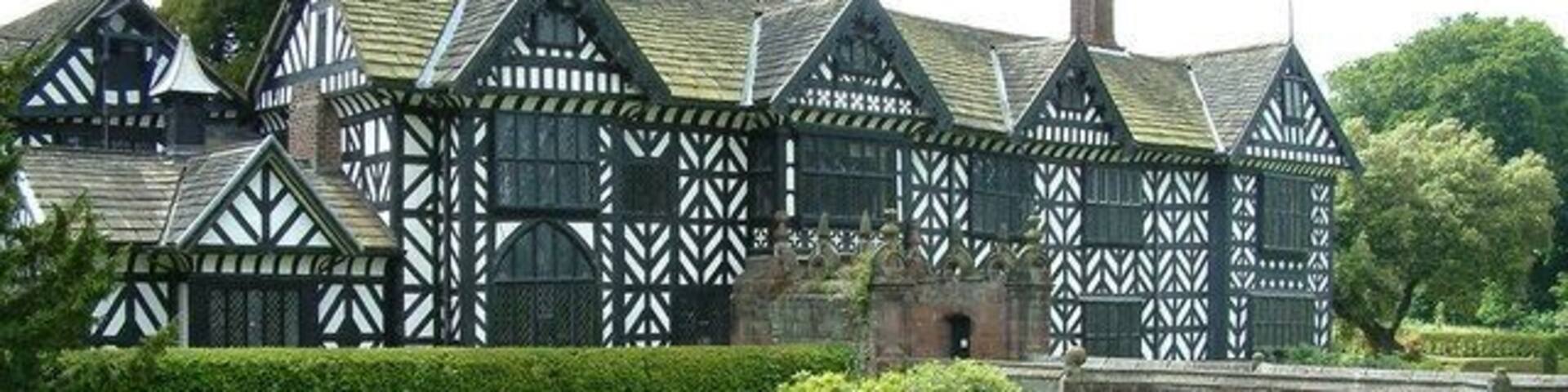 A Tudor-style building with white timber framing and a brick base, surrounded by lush greenery and a stone wall.