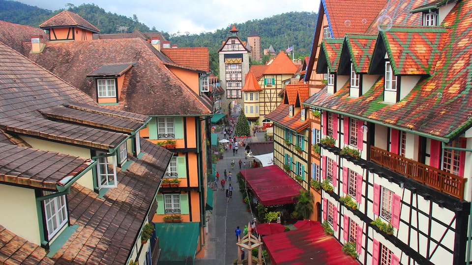 A narrow street lined with colorful half-timbered houses, a church steeple, and a mountain in the background.