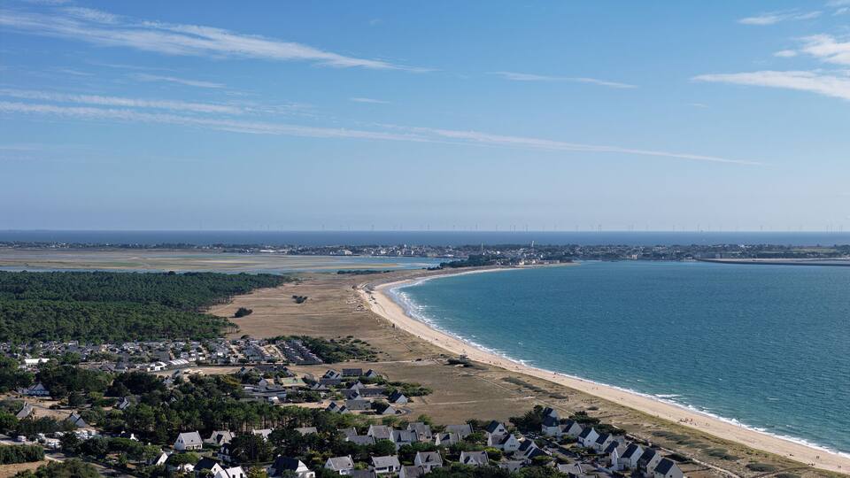 A coastal town with a beach, houses, and wind turbines in the distance.
