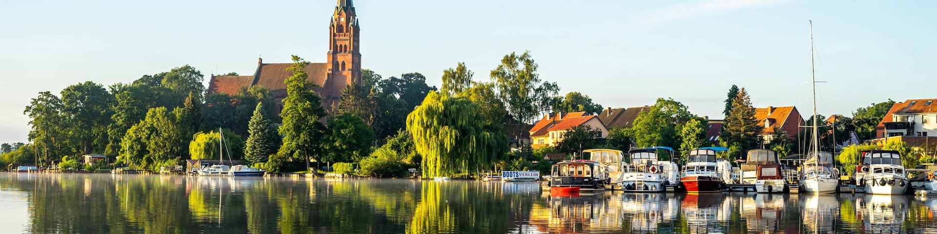 Un lago tranquilo con barcos, una iglesia con una aguja alta y edificios residenciales a lo largo de la orilla.