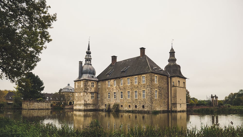 Un bâtiment historique en pierre, doté de plusieurs tours, dont le reflet se mire dans l’eau.