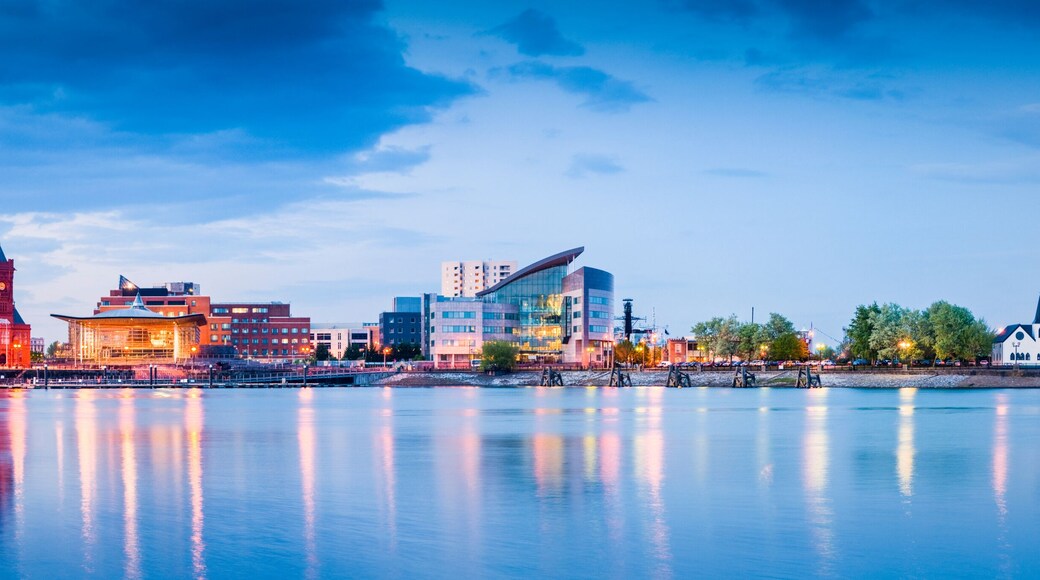 A waterfront cityscape with a mix of historic and modern architecture, including a prominent red building with a tower, a modern glass structure, and a large dome-like building.