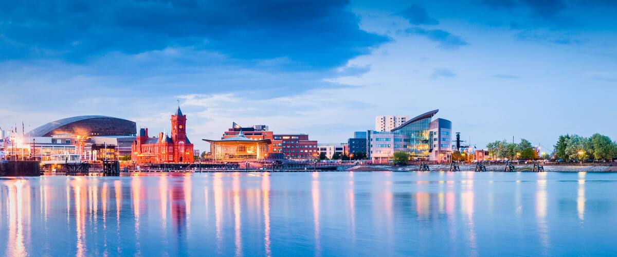 A waterfront cityscape with a mix of historic and modern architecture, including a prominent red building with a tower, a modern glass structure, and a large dome-like building.