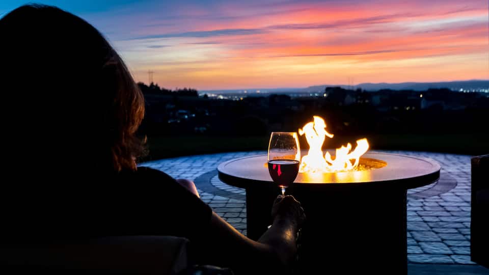 A person sitting by a fire pit with a glass of red wine, overlooking a sunset with a cityscape in the background.