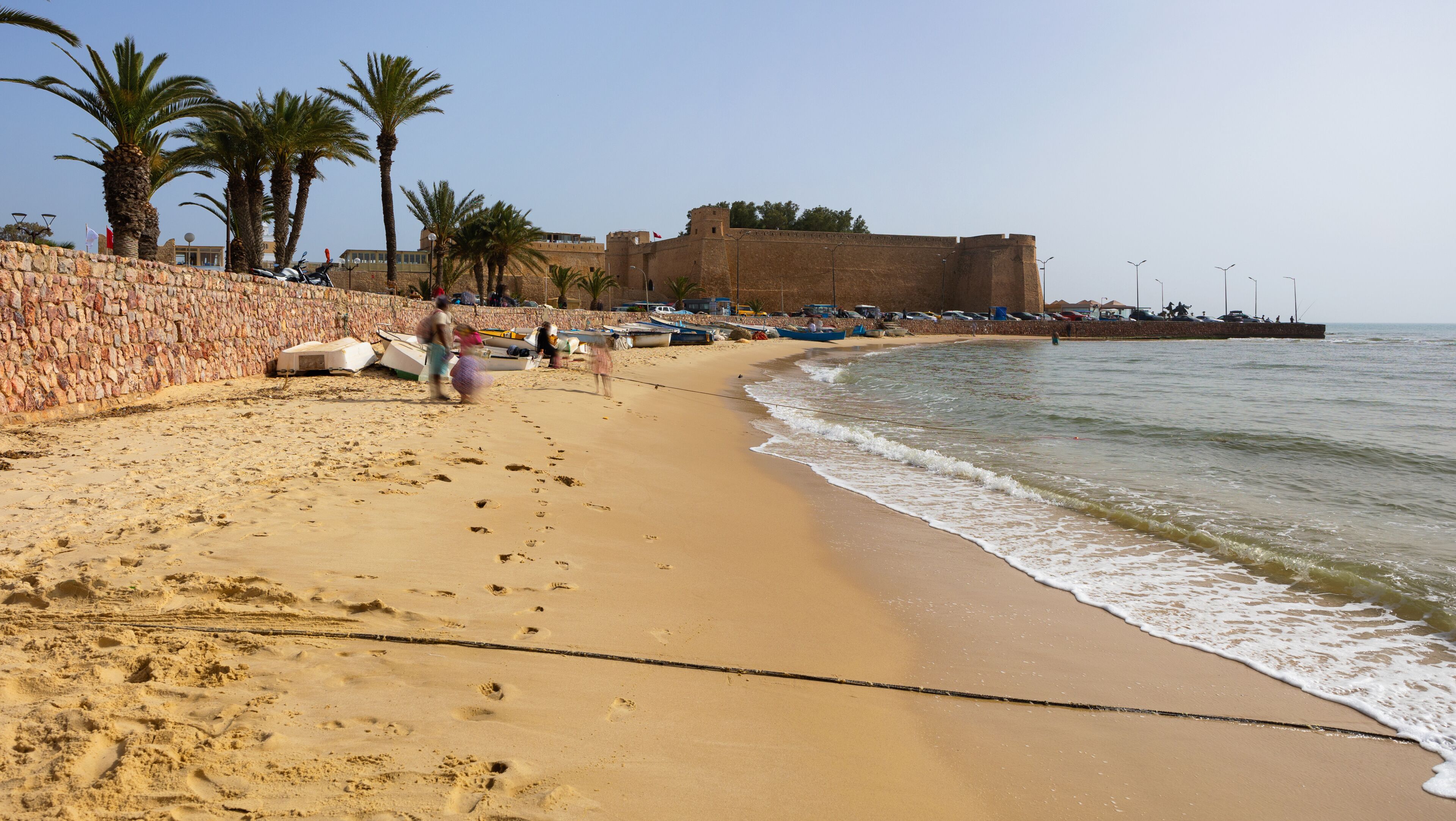 A sandy beach with palm trees, a stone wall, and a large fort-like structure by the water.