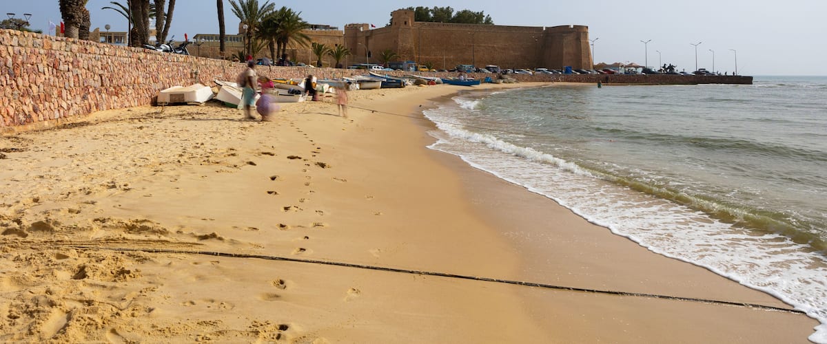 A sandy beach with palm trees, a stone wall, and a large fort-like structure by the water.
