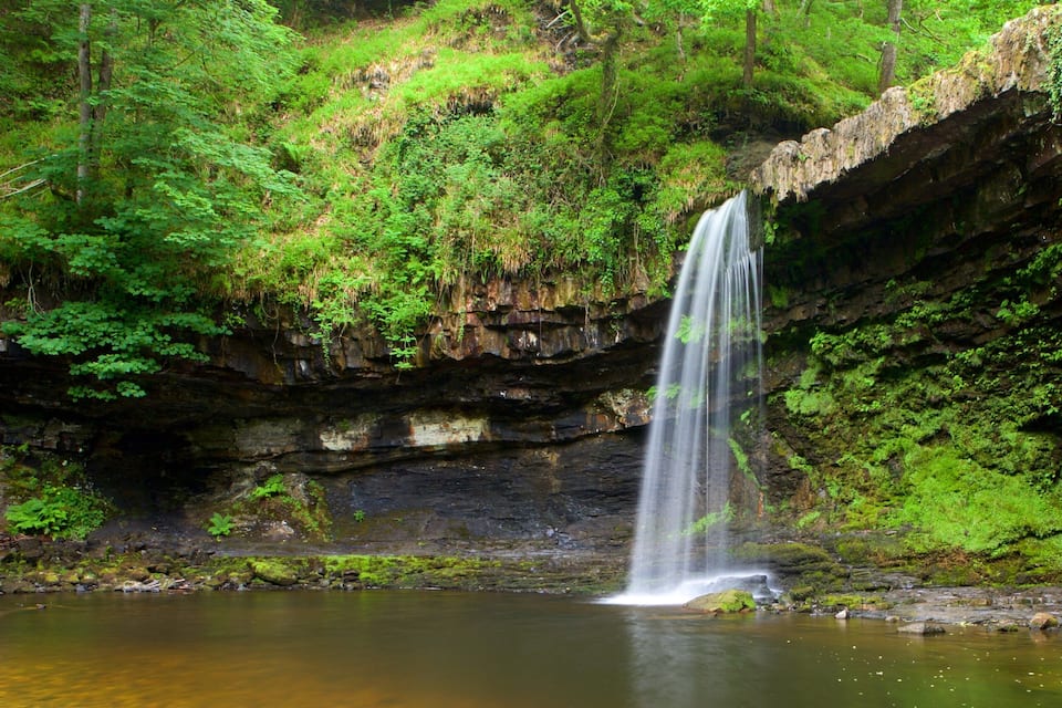 Uma cascata despencando em uma piscina calma, cercada por vegetação exuberante e paredões rochosos.