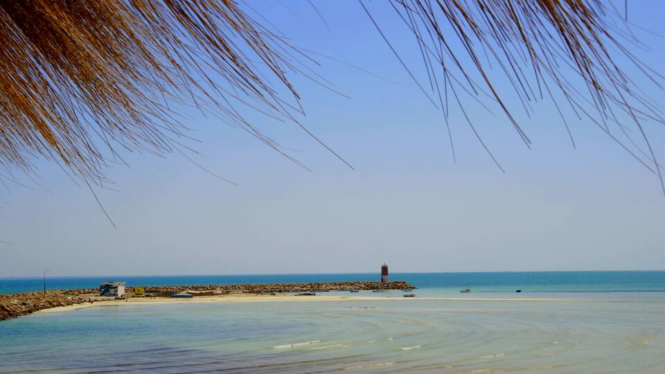 A beach with clear turquoise water, a rocky breakwater, and a lighthouse in the distance.
