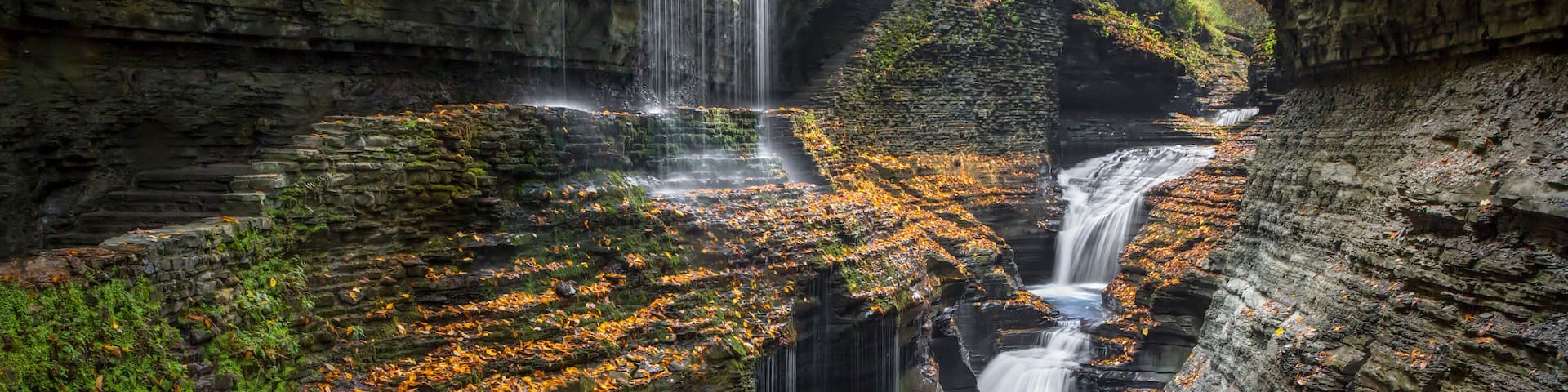 A multi-tiered waterfall cascading into a pool surrounded by moss-covered rocks and autumn leaves.