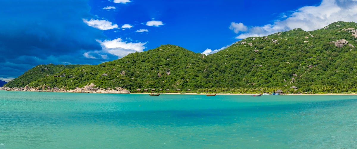 A tropical beach with clear turquoise waters, boats, and lush green mountains.