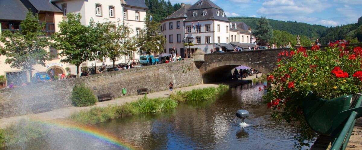 A river with a rainbow, a stone bridge, and buildings in the background.