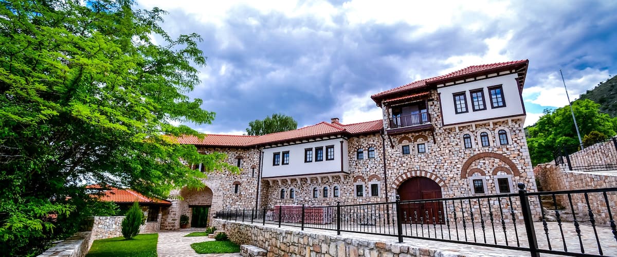 A historic stone building with a red-tiled roof, surrounded by a stone wall and a paved walkway.