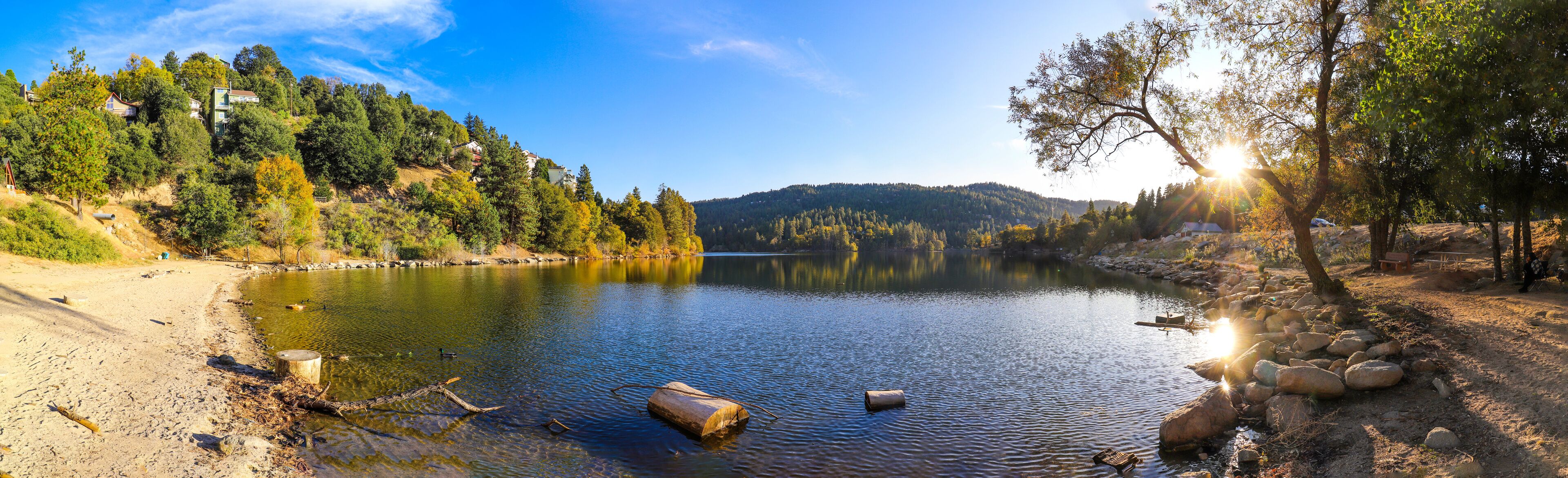 a breathtaking panoramic shot of the vast still blue lake waters with lush green and autumn colored trees reflecting off the lake with blue sky and mountain ranges at Lake Gregory Crestline California