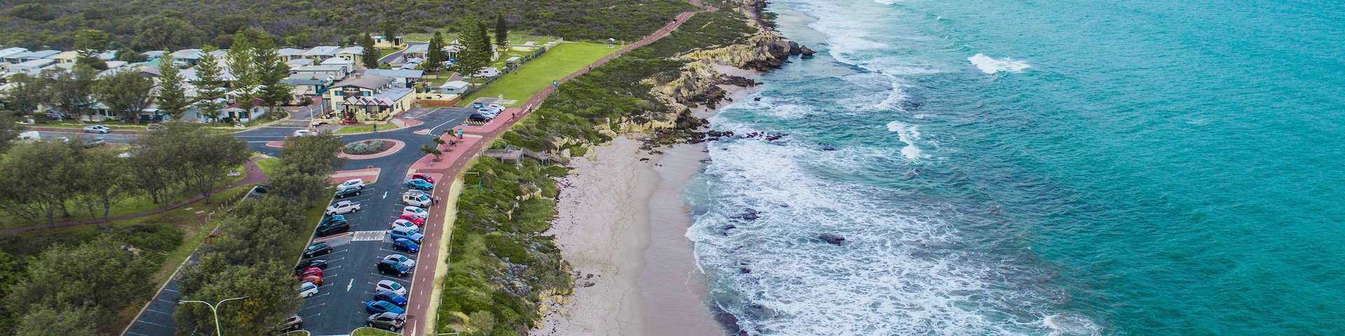 An aerial view of a coastal area with a beach, a parking lot, and a road leading to a residential area.