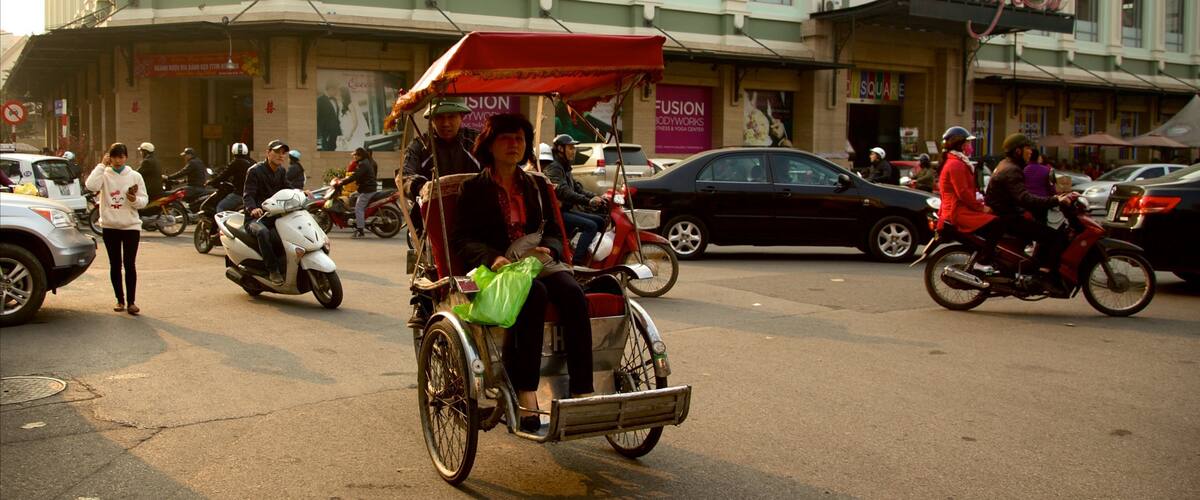 A busy street with a rickshaw, motorcycles, and cars. There are buildings with signs, including one that says "Fusion Body Works."