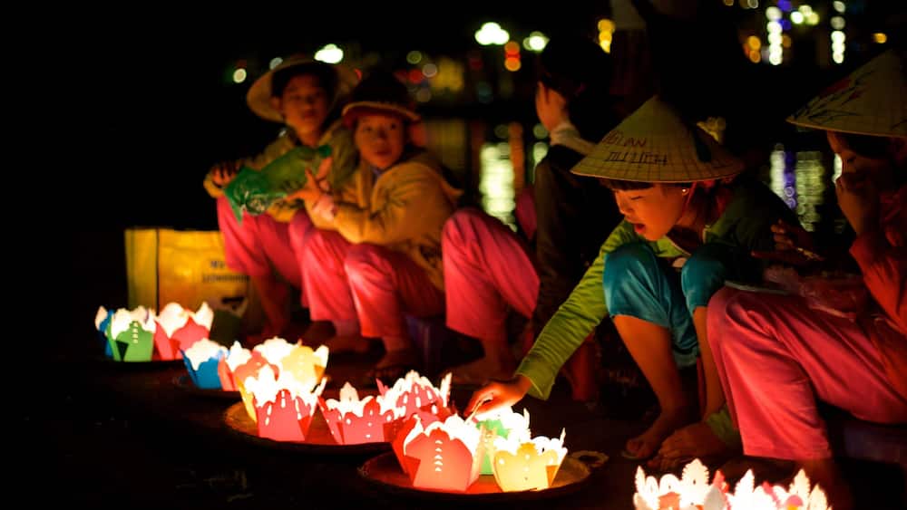 Des personnes portant des chapeaux traditionnels et des vêtements colorés sont assises autour d’une table, et des lanternes en papier illuminées sont disposées autour d’eux.