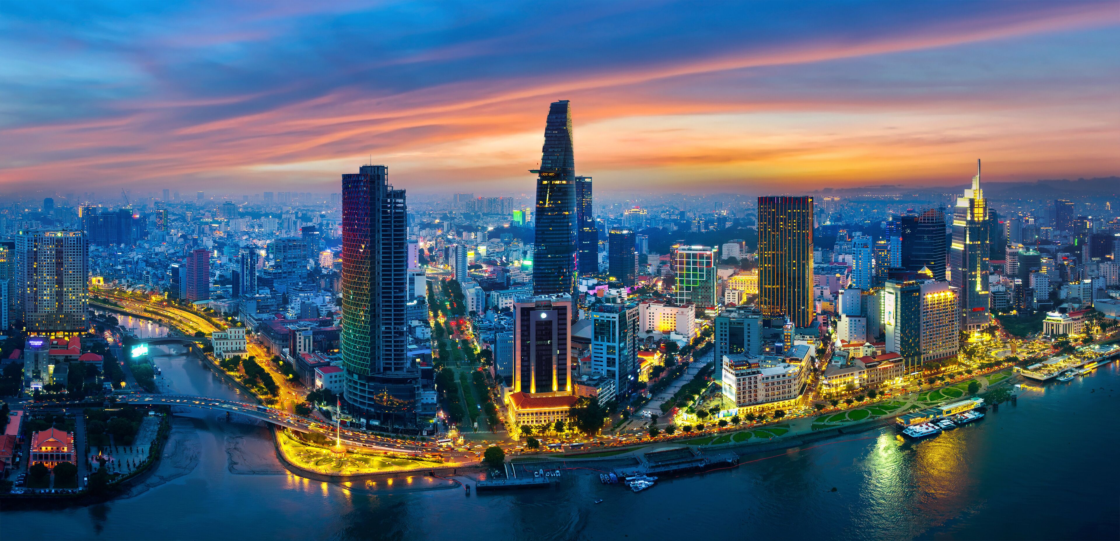 A cityscape with modern skyscrapers, a river, and a bridge at dusk.