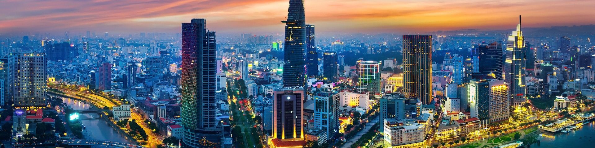 A cityscape with modern skyscrapers, a river, and a bridge at dusk.