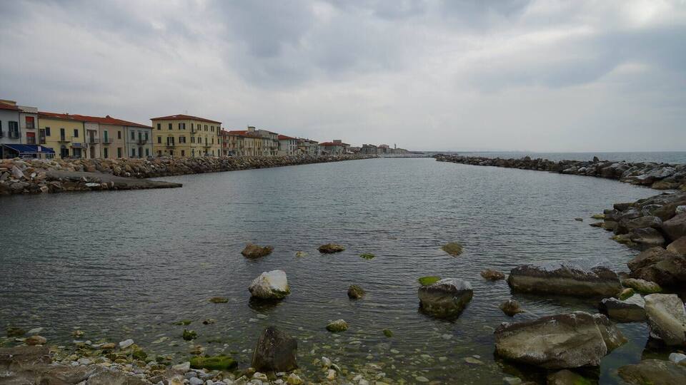 A coastal town with buildings along the shore, a rocky breakwater, and calm water.