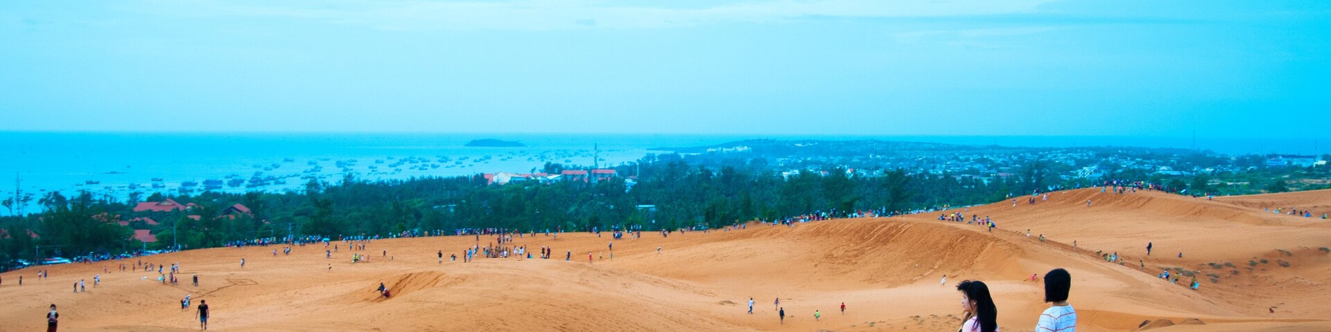 Paisaje arenoso con gente caminando y una vista distante del océano y la ciudad.