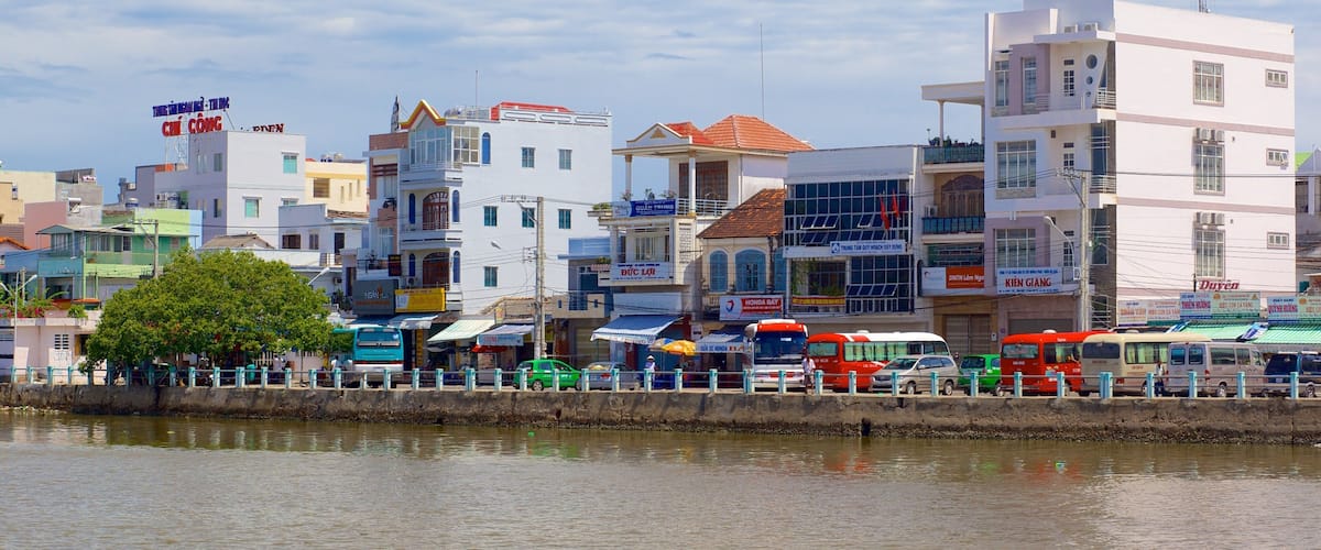 A riverside cityscape with various buildings, including a white building with a red roof and a blue sign.