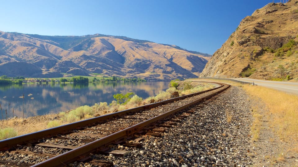 A railway track curving alongside a lake with mountains in the background.