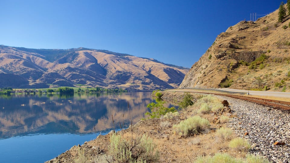 A lake with mountains in the background, a train track, and a clear sky.