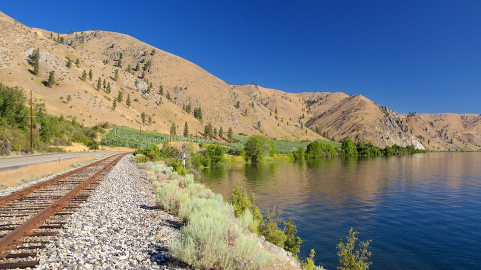 A railway track alongside a calm lake with mountains in the background.