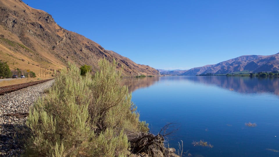 A calm lake with mountains in the background, a railway track on the left, and sparse vegetation in the foreground.
