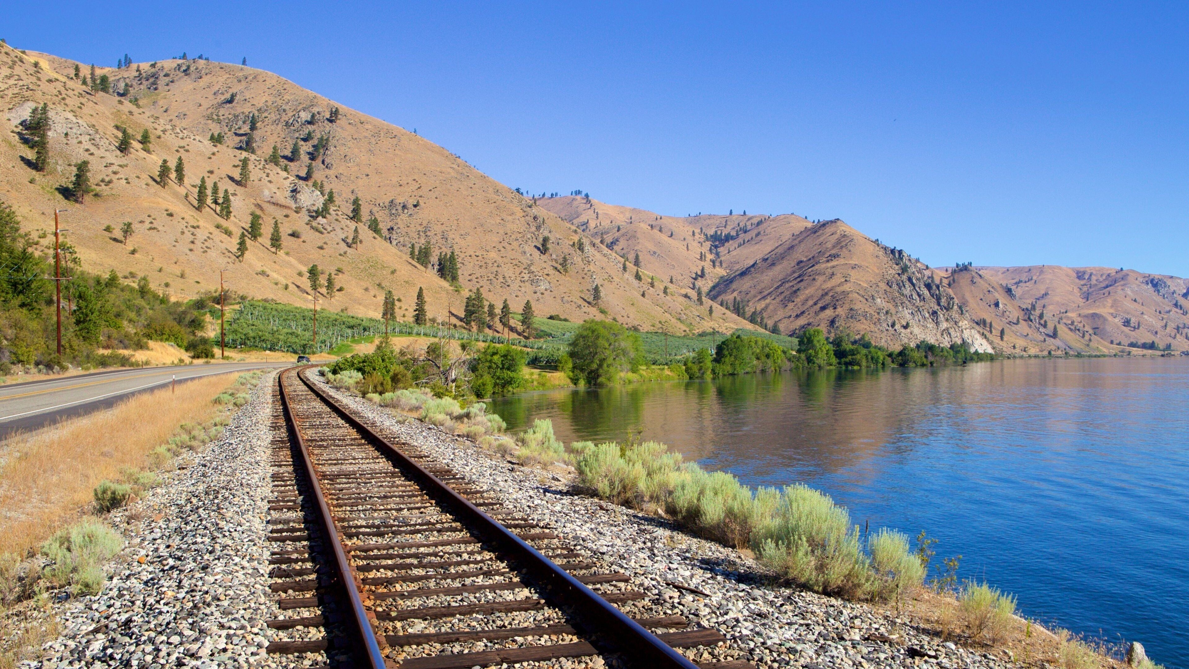 A railway track curving alongside a river, with mountains in the background.