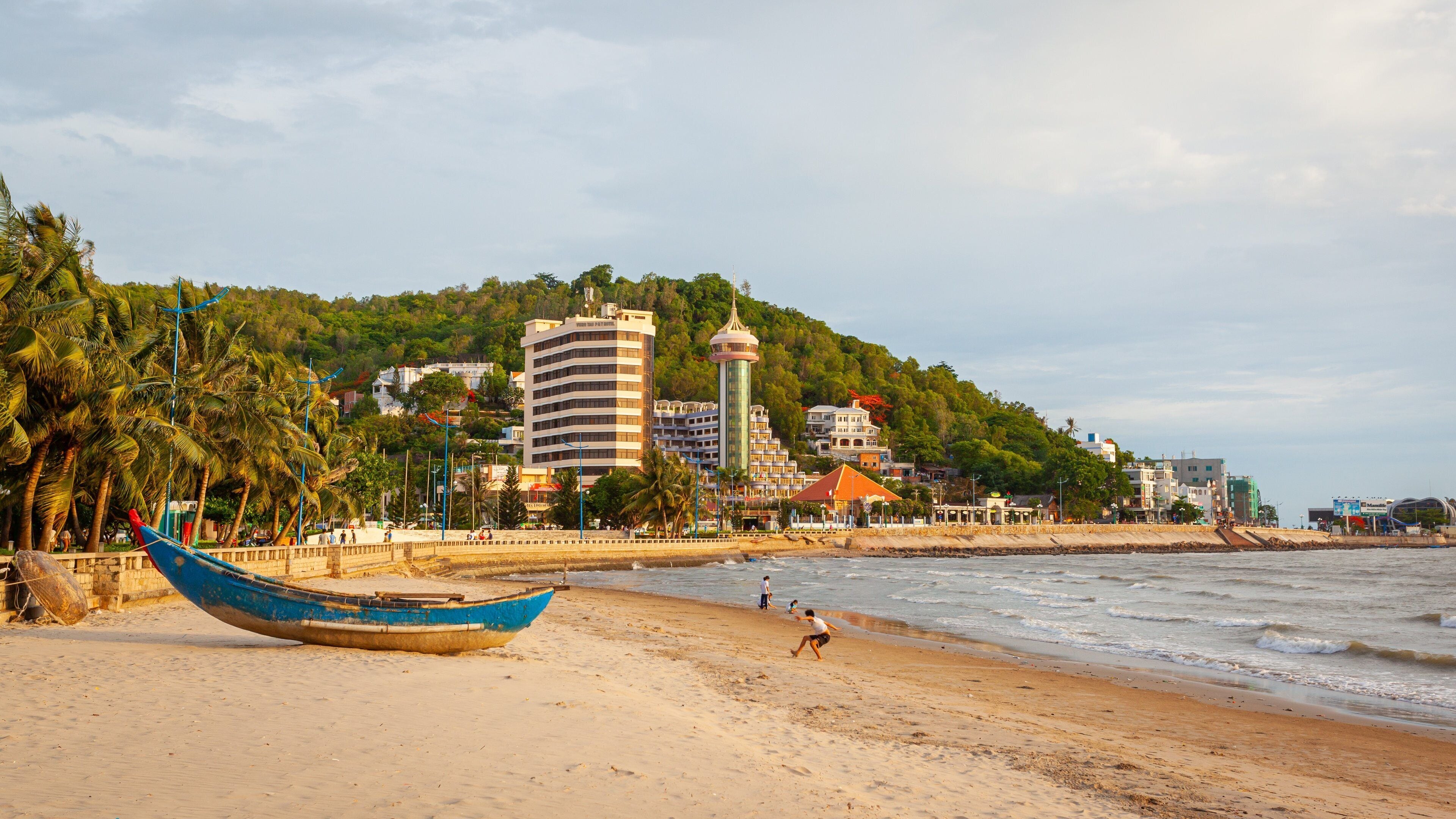 A beach with a blue boat, palm trees, and buildings on a hillside.