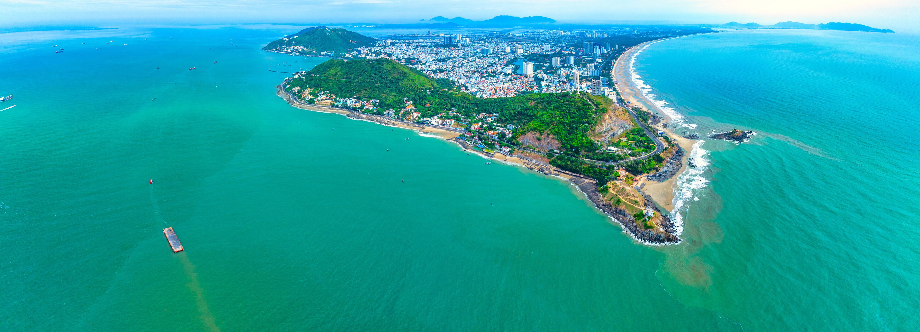 An aerial view of a coastal city with a prominent beach, dense urban area, and a green hill.