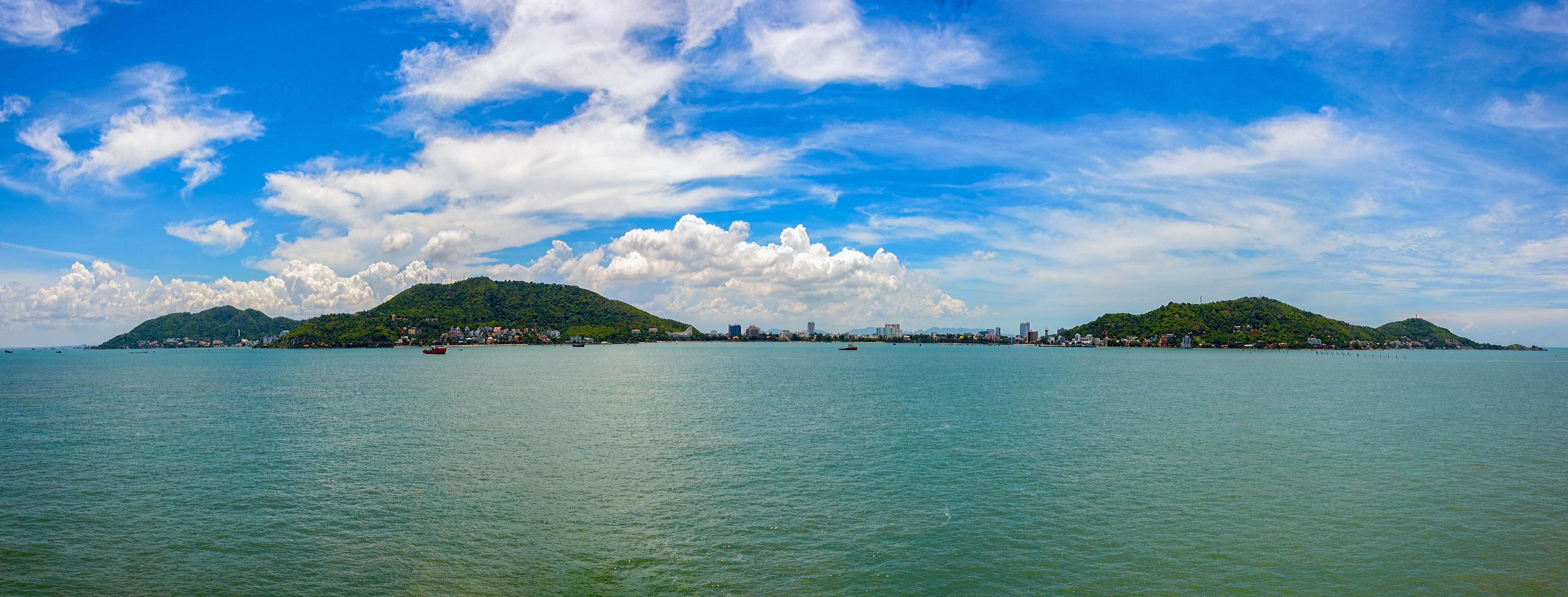 A coastal cityscape with two green hills, a clear blue sky, and calm sea.